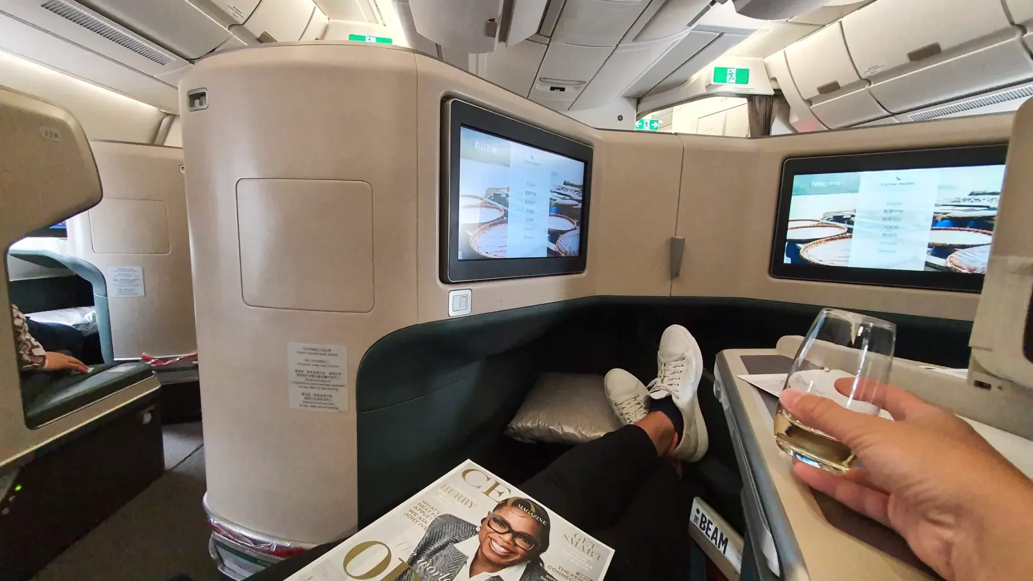 Inside a Business Class cabin: a passenger relaxing in a lie-flat suite, feet up with a drink in hand